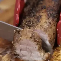 Un jugoso solomillo de cerdo al horno, recién cortado en una tabla de madera. Las pinzas rojas sujetan la carne mientras el cuchillo la lonchea.