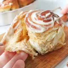 Un rollo de canela recién horneado, cubierto con glaseado cremoso, se desmenuza suavemente al ser tomado.