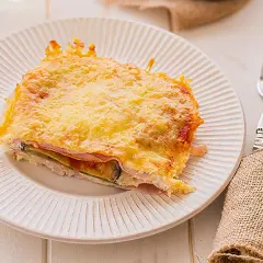 Porción de pastel de berenjenas gratinado con queso, servido en plato blanco con tenedor y servilleta de arpillera.