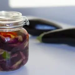 Tarro de berenjenas en escabeche con trozos de pimiento rojo y hierbas. Dos berenjenas frescas en el fondo.