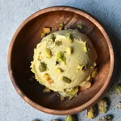 Cremoso helado de pistacho servido en un cuenco de madera, adornado con trozos de pistacho y acompañado de más pistachos enteros esparcidos alrededor.
