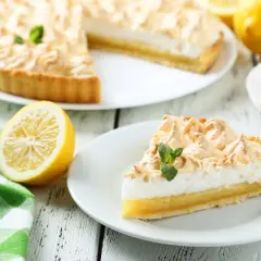 Una rebanada de tarta de limón con merengue, decorada con menta, junto a un limón y una taza de café.