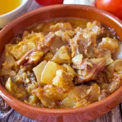 Guiso de cap i pota en cazuela de barro, con patatas y trozos de carne, rodeado de ingredientes como aceite, pimentón y tomates.