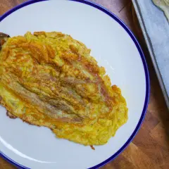 Tortilla de berenjena dorada y jugosa, servida en plato blanco con borde azul. Al lado, una bandeja con papel de horno y más berenjena.