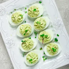 Deliciosos huevos rellenos de aguacate, decorados con brotes verdes y especias, servidos en una bandeja blanca con relieve floral.