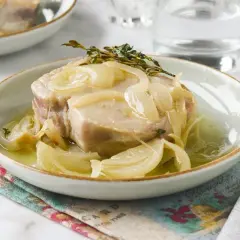 Un trozo de atún en escabeche con cebolla y tomillo, servido en un plato blanco con un tenedor al lado.