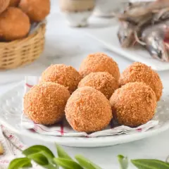 Croquetas de gambas doradas y crujientes en un plato blanco, con más en una cesta de mimbre al fondo y gambas frescas a la derecha.