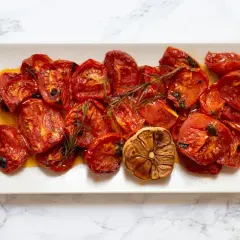 Tomates al horno, jugosos y caramelizados, con ajo asado y romero, servidos en una bandeja blanca sobre mármol.