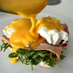 Un delicioso uovo alla benedict, con huevo escalfado, jamón, rúcula y salsa holandesa cremosa vertiéndose sobre él.