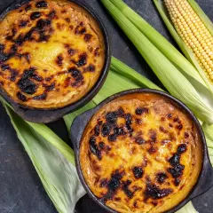 Dos cazuelas de pastel de choclo recién horneado, con su dorada y caramelizada cubierta, junto a mazorcas de maíz fresco.