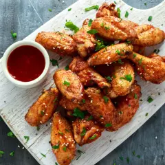 Alitas de pollo al horno crujientes, doradas y espolvoreadas con sésamo y perejil, servidas con salsa roja en un cuenco.