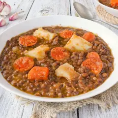 Un plato humeante de lentejas con chorizo, patatas y zanahoria, servido en un cuenco blanco sobre una mesa de madera.