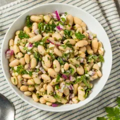 Deliciosa ensalada de judías blancas con cebolla morada y perejil fresco, servida en un cuenco blanco sobre un mantel a rayas.
