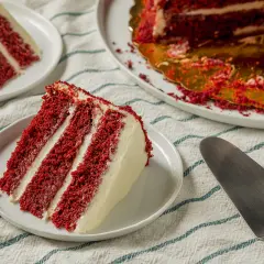 Porción de pastel red velvet con capas de bizcocho rojo y crema blanca. Otro trozo y el pastel entero en el fondo.