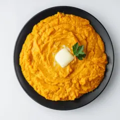 Un cremoso puré de boniato, coronado con una pastilla de mantequilla y perejil fresco, listo para disfrutar.