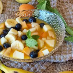 Deliciosa macedonia de frutas con plátano, arándanos, melocotón y manzana en un cuenco de cristal, decorada con menta.
