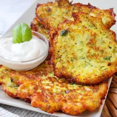 Tortitas de calabacín doradas y crujientes, servidas con una cremosa salsa y decoradas con una hoja de albahaca.