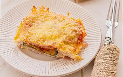 Porción de pastel de berenjenas gratinado con queso, servido en plato blanco con tenedor y servilleta de arpillera.