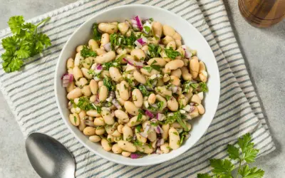 Deliciosa ensalada de judías blancas con cebolla morada y perejil fresco, servida en un cuenco blanco sobre un mantel a rayas.