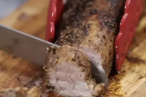 Un jugoso solomillo de cerdo al horno, recién cortado en una tabla de madera. Las pinzas rojas sujetan la carne mientras el cuchillo la lonchea.