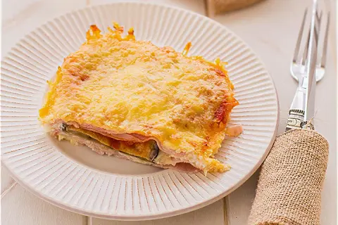 Porción de pastel de berenjenas gratinado con queso, servido en plato blanco con tenedor y servilleta de arpillera.