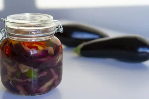 Tarro de berenjenas en escabeche con trozos de pimiento rojo y hierbas. Dos berenjenas frescas en el fondo.
