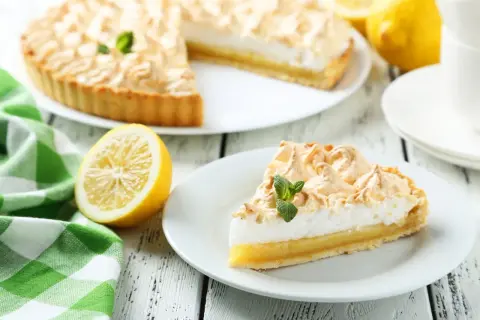 Una rebanada de tarta de limón con merengue, decorada con menta, junto a un limón y una taza de café.