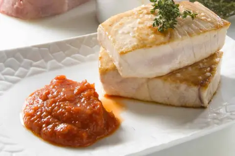 Dos trozos de atún a la plancha, dorados y jugosos, apilados sobre un plato blanco con una porción de salsa de tomate y una ramita de tomillo.