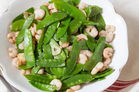 Plato de tirabeques salteados con gambas y ajo. Los tirabeques, crujientes y verdes, se mezclan con las gambas rosadas y láminas de ajo.