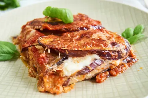 Un trozo de parmigiana di melanzane, con capas de berenjena, queso fundido y salsa de tomate, adornado con hojas de albahaca fresca.
