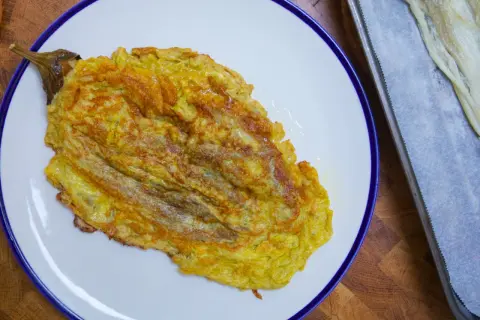 Tortilla de berenjena dorada y jugosa, servida en plato blanco con borde azul. Al lado, una bandeja con papel de horno y más berenjena.