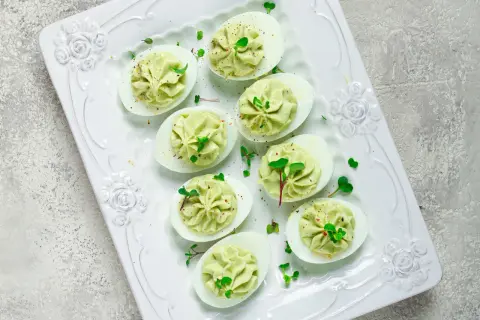 Deliciosos huevos rellenos de aguacate, decorados con brotes verdes y especias, servidos en una bandeja blanca con relieve floral.
