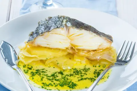 Merluza al horno con patatas, jugosa y dorada, servida sobre una cama de patatas y cebolla caramelizada, bañada en aceite de oliva y perejil picado.