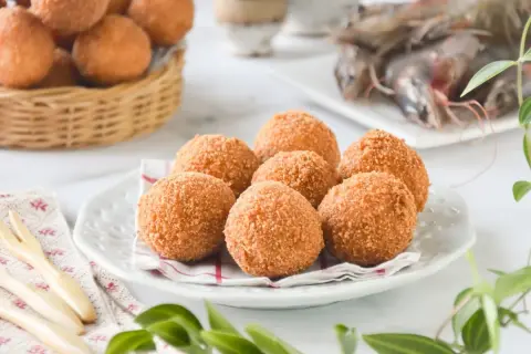 Croquetas de gambas doradas y crujientes en un plato blanco, con más en una cesta de mimbre al fondo y gambas frescas a la derecha.