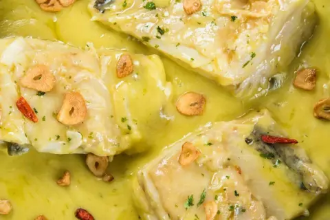 Merluza al pil pil, jugosos trozos de pescado blanco en una salsa cremosa con láminas de ajo crujiente y guindilla.