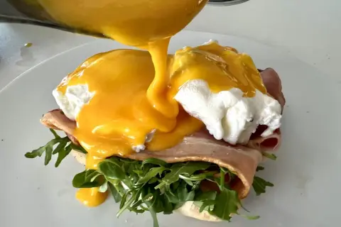 Un delicioso uovo alla benedict, con huevo escalfado, jamón, rúcula y salsa holandesa cremosa vertiéndose sobre él.