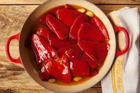 Pimientos del piquillo asados en una cazuela roja, con ajos y un paño de cocina.