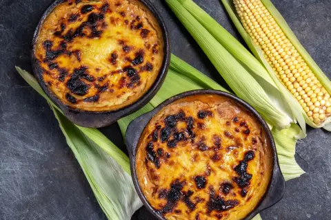 Dos cazuelas de pastel de choclo recién horneado, con su dorada y caramelizada cubierta, junto a mazorcas de maíz fresco.
