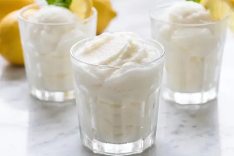 Tres vasos de sorbete de limón, refrescante y cremoso, con limones amarillos y hojas de menta al fondo.