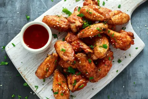 Alitas de pollo al horno crujientes, doradas y espolvoreadas con sésamo y perejil, servidas con salsa roja en un cuenco.