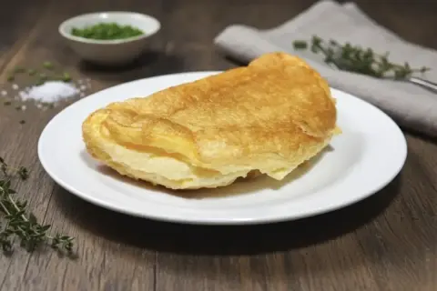 Tortilla francesa dorada y esponjosa servida en plato blanco, con hierbas y sal alrededor.