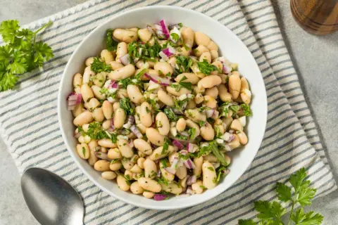 Deliciosa ensalada de judías blancas con cebolla morada y perejil fresco, servida en un cuenco blanco sobre un mantel a rayas.