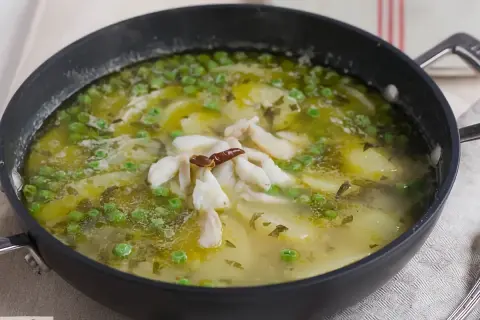Patatas en salsa verde con trozos de pescado blanco y guisantes, servido en una sartén negra.