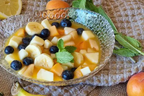 Deliciosa macedonia de frutas con plátano, arándanos, melocotón y manzana en un cuenco de cristal, decorada con menta.
