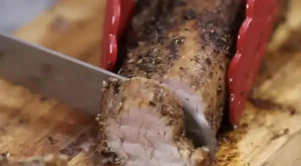 Un jugoso solomillo de cerdo al horno, recién cortado en una tabla de madera. Las pinzas rojas sujetan la carne mientras el cuchillo la lonchea.
