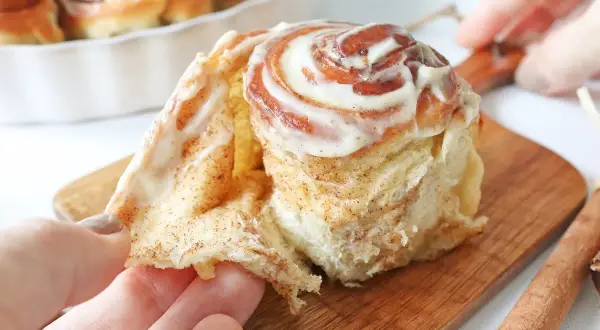 Un rollo de canela recién horneado, cubierto con glaseado cremoso, se desmenuza suavemente al ser tomado.