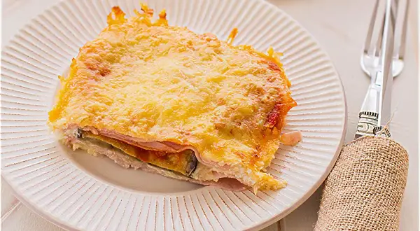 Porción de pastel de berenjenas gratinado con queso, servido en plato blanco con tenedor y servilleta de arpillera.