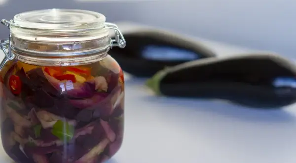 Tarro de berenjenas en escabeche con trozos de pimiento rojo y hierbas. Dos berenjenas frescas en el fondo.