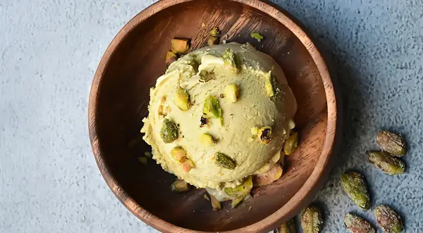 Cremoso helado de pistacho servido en un cuenco de madera, adornado con trozos de pistacho y acompañado de más pistachos enteros esparcidos alrededor.