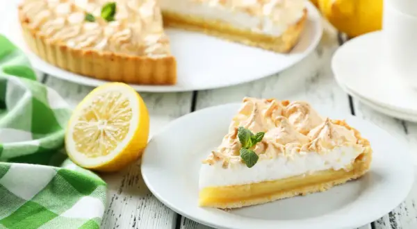 Una rebanada de tarta de limón con merengue, decorada con menta, junto a un limón y una taza de café.